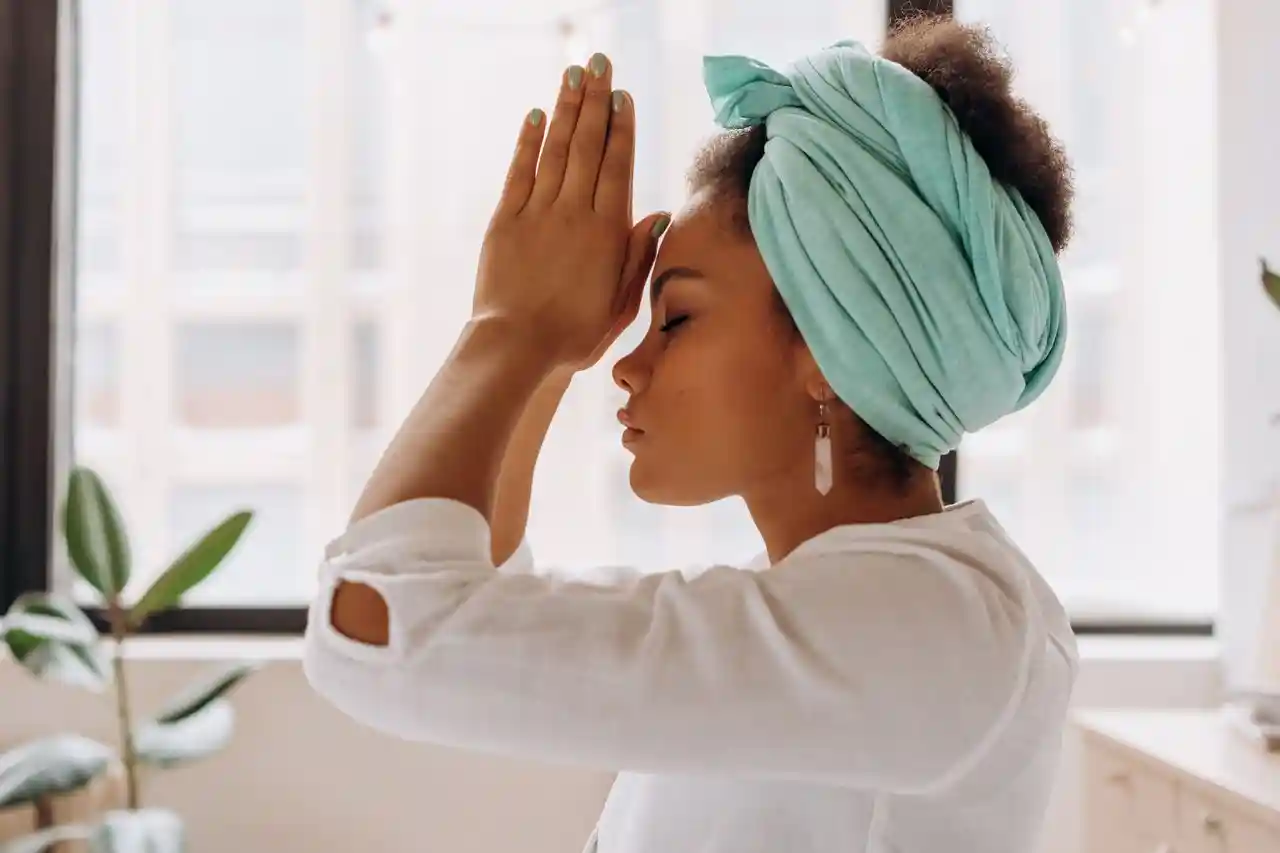 Woman in a turquoise head wrap with hands in prayer position, eyes closed in quiet reflection
