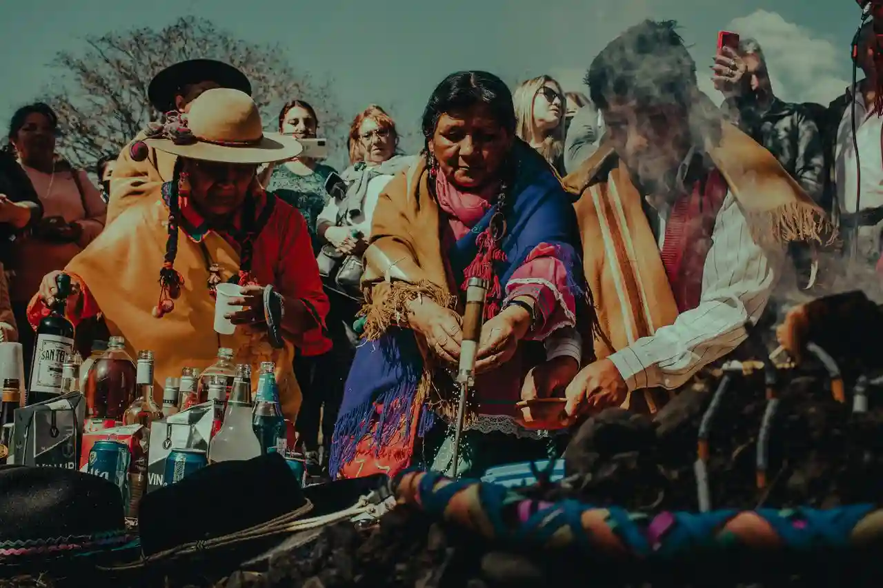 Women in Andean dress place offerings around a smoking ceremonial fire while onlookers watch.